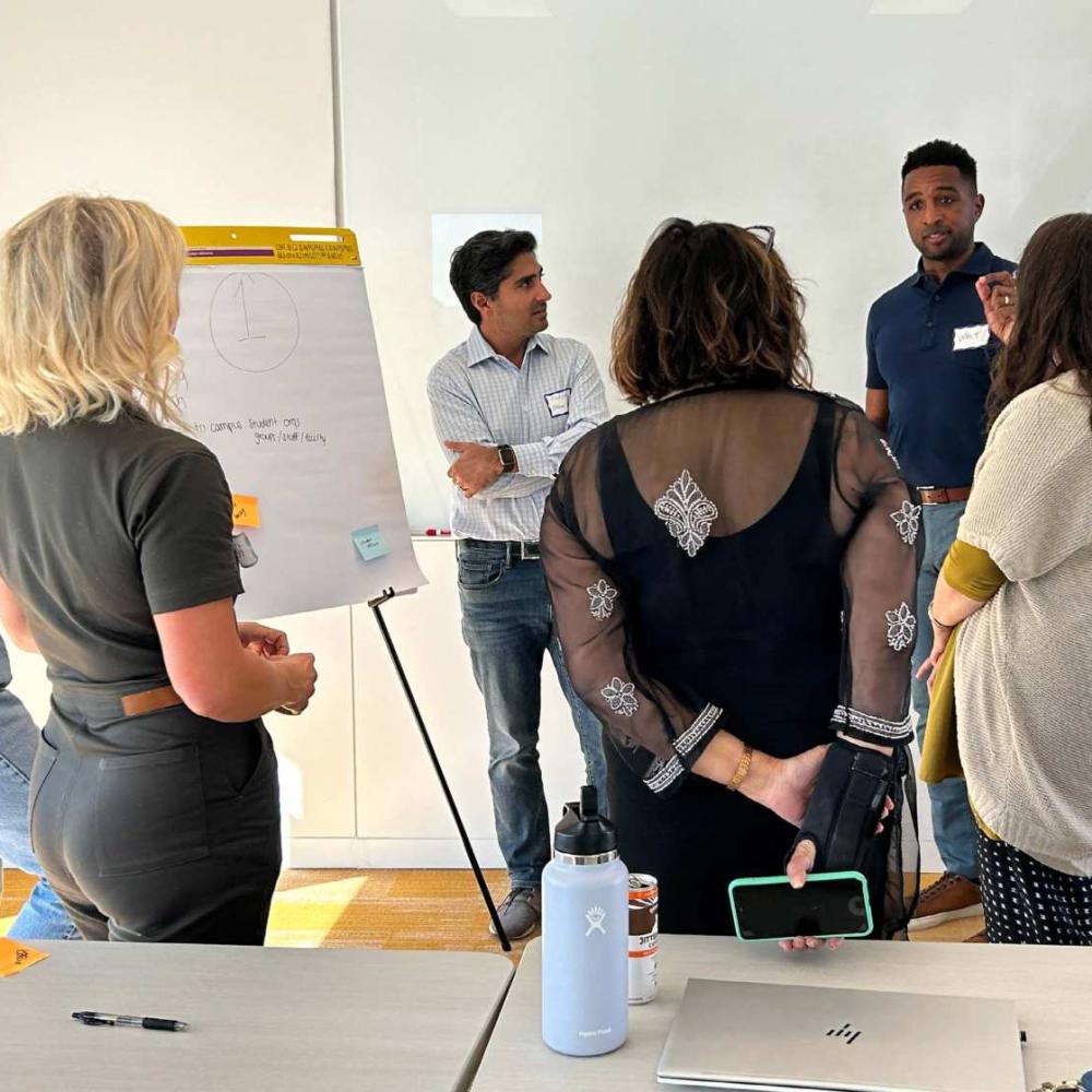 Group of four people looking and listening to presenter while standing in front of easel with notes 